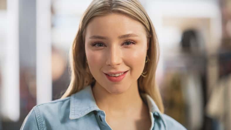 Portrait of Beautiful Young Woman with Light Blonde Hair and Green Eyes Wearing Jeans Shirt Looking Up to the Camera and Smiling. Successful Woman Posing for Camera. Blurred Clothing Store Background.