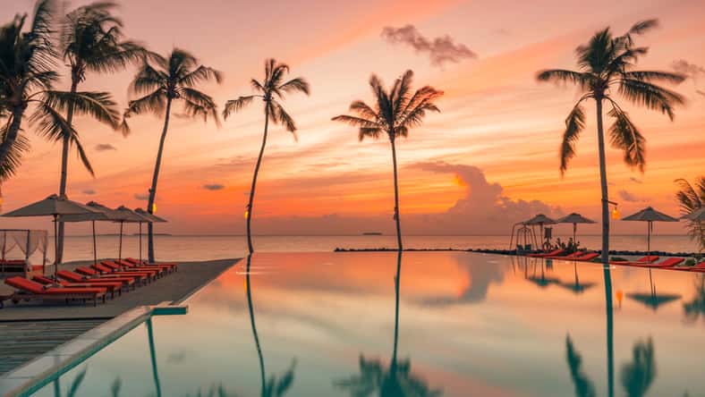 Fantastic poolside, sunset sky, palm trees reflection. Luxury tropical beach landscape, infinity swimming pool, deck chairs and loungers under umbrellas amazing scenic. Vacation resort hotel landscape