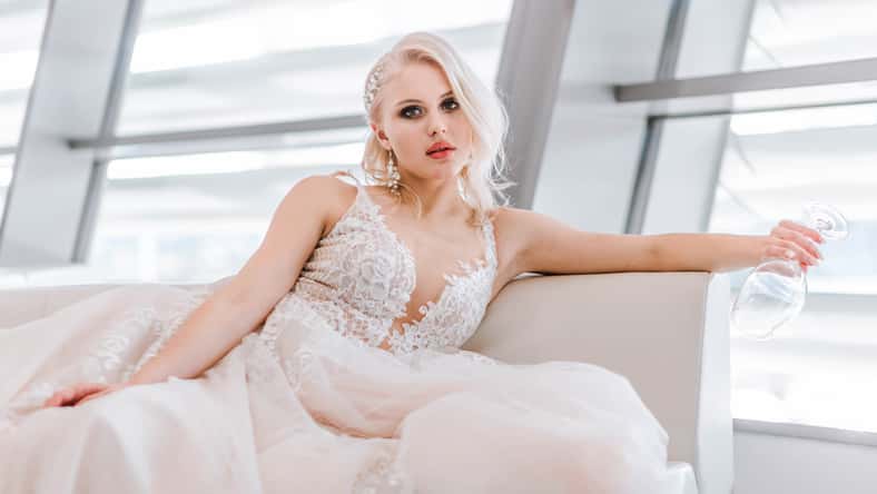An attractive pensive blonde woman with a creative hairstyle for short hair is sitting on a white sofa in the lobby of the hotel. Art photography. White long wedding dress of the bride.