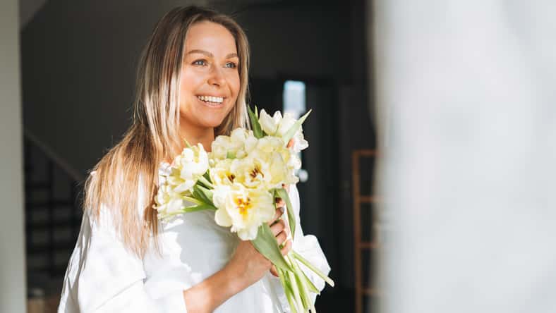 Young beautiful smiling woman forty year with blonde long hair in white shirt with bouquet of yellow flowers in hands near window in bright interior at home