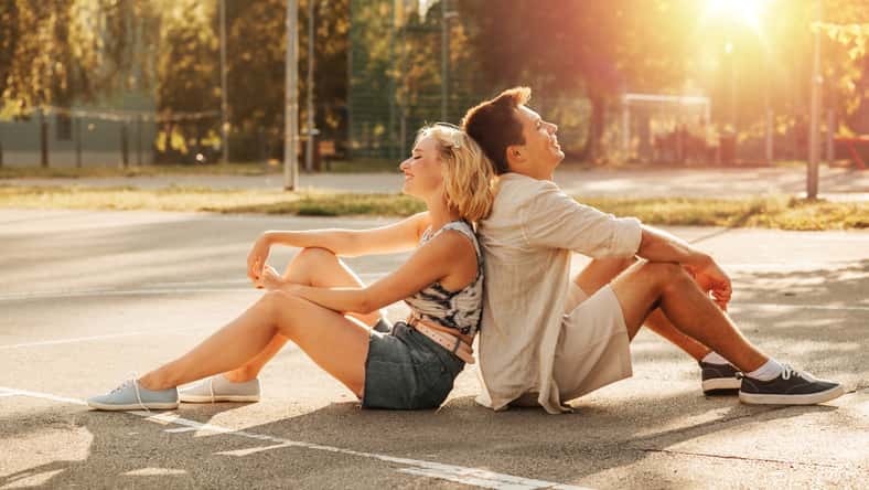 summer holidays and people concept - happy young couple sitting back to back on basketball playground
