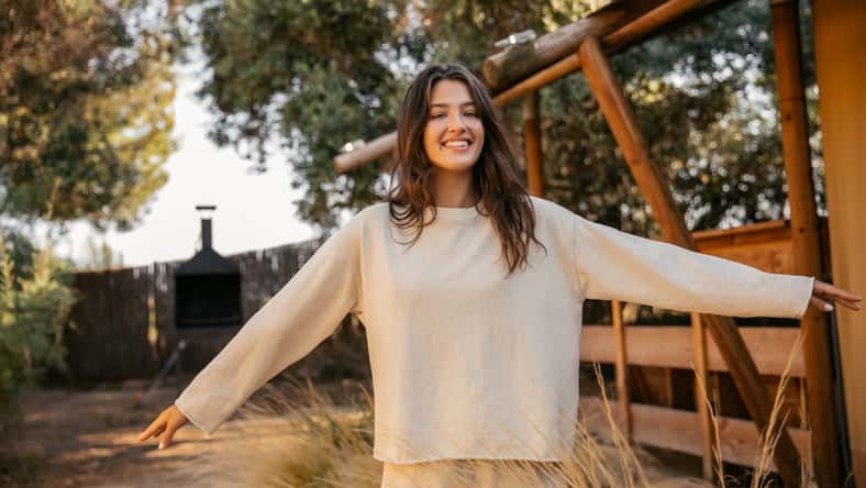 Positive young caucasian girl waving her arms walking in countryside during day. Brunette smiles broadly, wears spring clothes. Nature, summer holidays concept