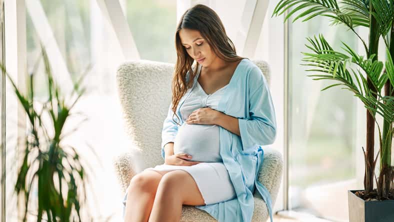 Pregnant happy woman touching her belly rest on chair at modern home. Tender mood photo of healthy pregnancy.
