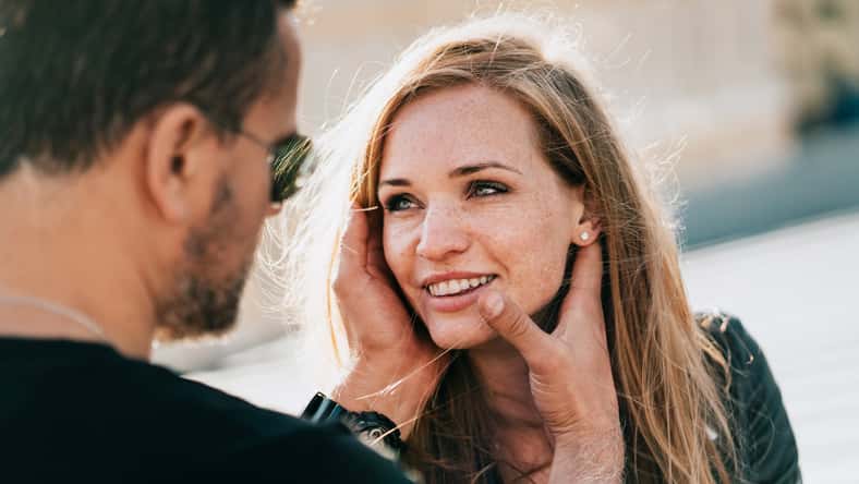 Close up of your adult American couple in black clothes looking at each other. Redhead cheerful caucasian girl looks tenderly at boyfriend. Romantic man touching wife’s face, date, relationship.