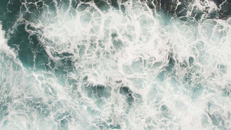 white sea foam on the ocean surface is agitated salt water from crashing waves and a strong surf, mixing currents and undertow swirl in the deep blue ocean during typhoon storms interrupt shipping