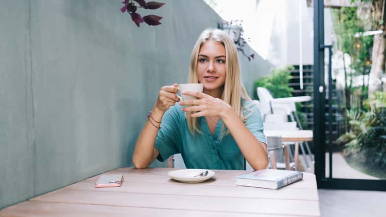 Carefree Caucasian woman with cup of hot caffeine beverage dreaming about leisure spending and positive youth lifestyle, happy blonde female customer resting in outdoors coffee shop enjoying tea time