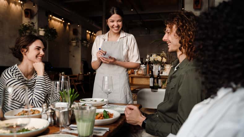 Young smiling waitress wearing apron serving group of cheerful young friends in restaurant