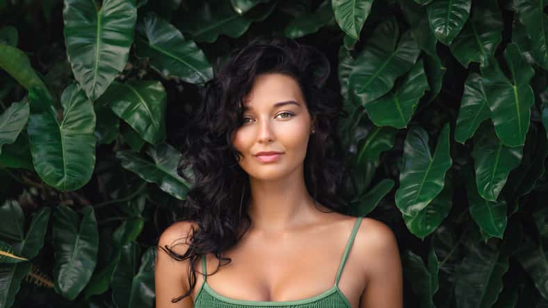 Natural beauty sport portrait woman with slim tanned body in stylish green sporty top posing near the wall of green exotic foliage and tropical leaves. High quality advertisement photo