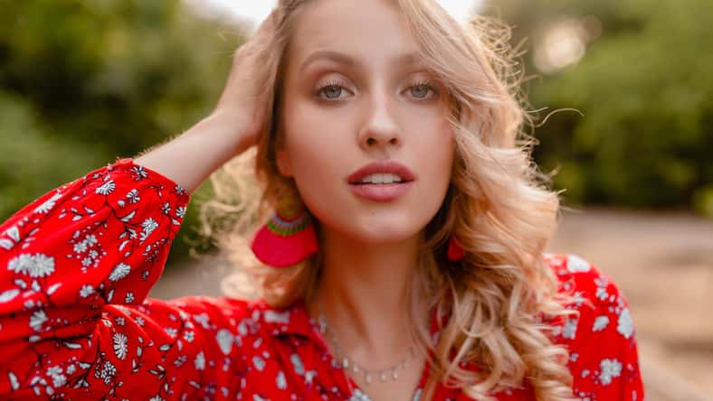 portrait of attractive stylish blond smiling woman in red blouse summer fashion outfit in park boho style wearing earrings smiling white teeth