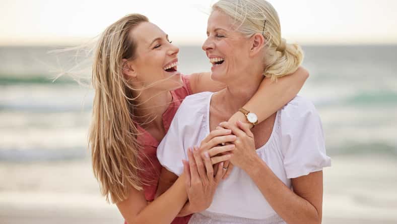 Smile, beach and senior mom with daughter for bonding holiday, love and embrace on weekend travel. Nature, mother and woman laughing on ocean adventure together with hug, fun and island vacation