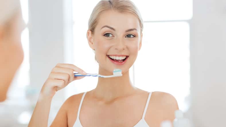 smiling young woman with toothbrush cleaning teeth and looking to mirror at home bathroom