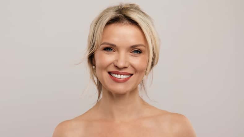 Portrait of cheerful attractive half-naked middle aged blonde woman smiling at camera, posing on grey studio background, demonstrating glowing perfect skin, copy space. Cosmetology concept. High quality photo