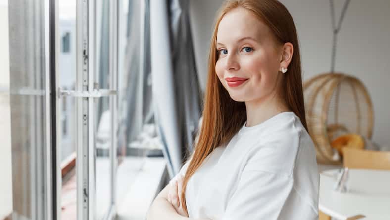 Side view of cute young girl in white t-shirt standing next to big window with folded arms, looking at camera with smile, youthful female student having rest after finishing homework