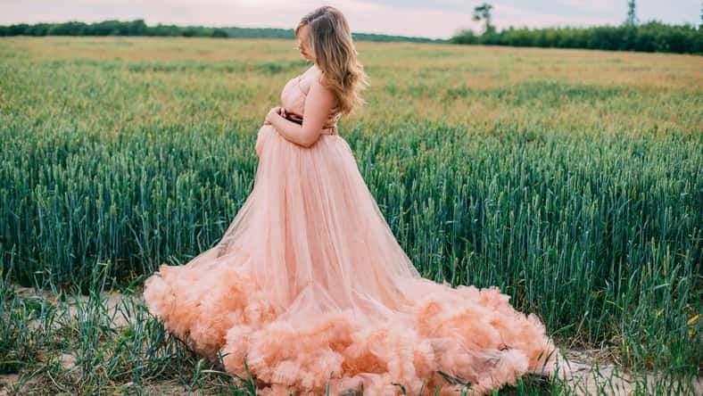 Young happy pregnant woman in peach color cloud dress relaxing and enjoying life in nature. Outdoors on the nature. Pretty pregnant woman holding her tummy. Cloudy dress. Blues sky. Wheat green field.