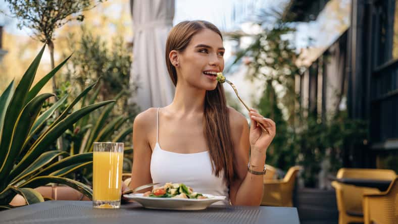 Eating food that is on the plate. Beautiful young woman is outdoors of the restaurant by the table.