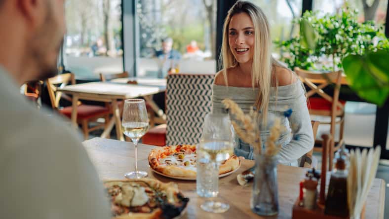 Young woman smiling at her boyfriend while having lunch at a pizzeria, enjoying pizza and white wine