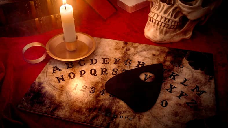 Una guarida de brujas, adivino y concepto de magia negra con un tablero de espíritu parlante iluminado por una vela rodeada de una baraja de cartas, tres libros y un cráneo envuelto con humo misterioso y turbio