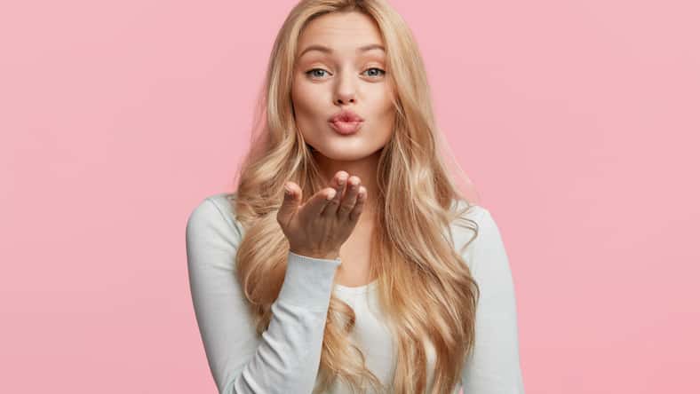 Pretty blonde woman blows passionate kiss into camera, expresses her love, isolated over pink background. Adorable young female model makes air kiss. People, body language and beauty concept