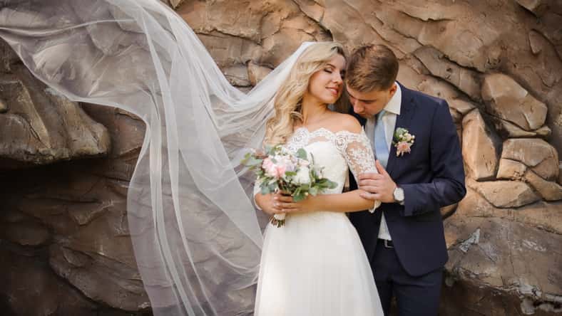 Portrait of a young bridegroom embracing a lovely blonde bride holding a bouquet of flowers on the background of wild stone