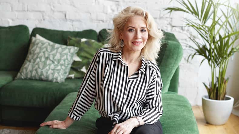 Indoor portrait of luxurious glamourous attractive European woman in her sixties with blonde hair and fit body looking at camera with pleased friendly smile, sitting on comfortable green couch