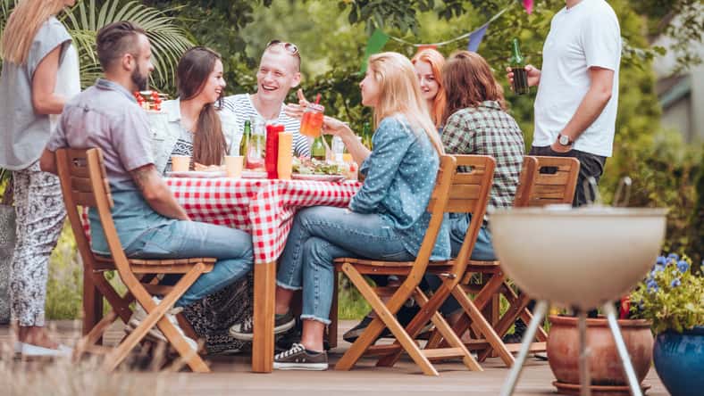 La gente se reunió alrededor de una mesa de madera, comiendo, bebiendo y divirtiéndose durante una fiesta de parrilla en la terraza.