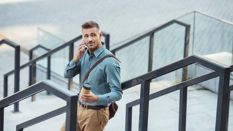 smiling middle aged man talking by smartphone hand holding paper cup while standing on stairs