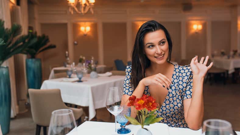 Happy brunette young elegant woman wearing dress sitting in the restaurant waving her hand to her friend. Caucasian female posing in cafeteria and waiting her family for a dinner. Copy space for text