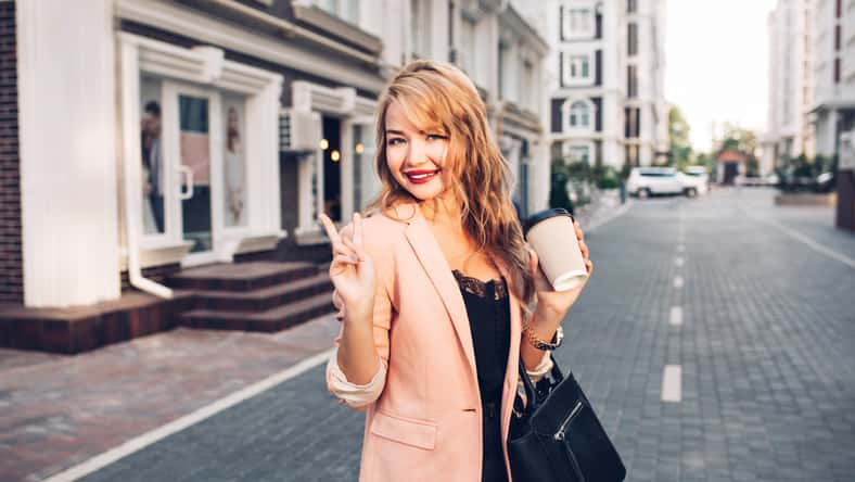 Portrait fashionable blonde woman with long hair walking in coral jacket on street. She holds a cup of coffee, smiling to camera