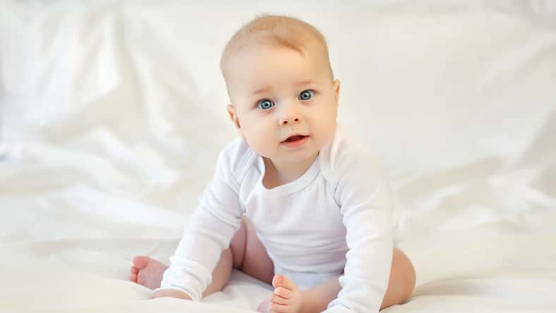 Charming blue-eyed 9 month old baby in white clothes sitting on the bed