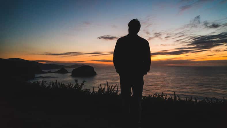 Back of a young traveler standing on on the edge of a cliff by the ocean coast watching a beautiful sunset. Freedom and wanderlust concept