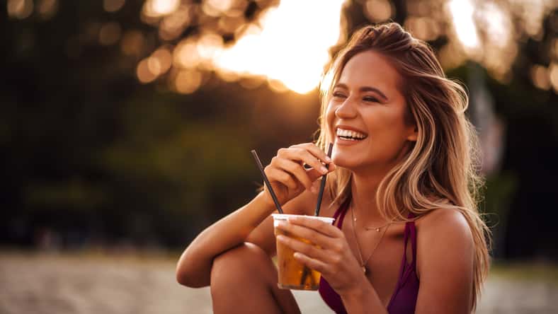 Close-up portrait of a beautiful happy young woman drinking on a beach. Summer vacation concept.
