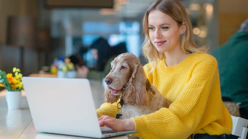 Young caucasian blonde freelancer girl holding her cocker spaniel dog on her knees in co working space and using laptop while working or studying.