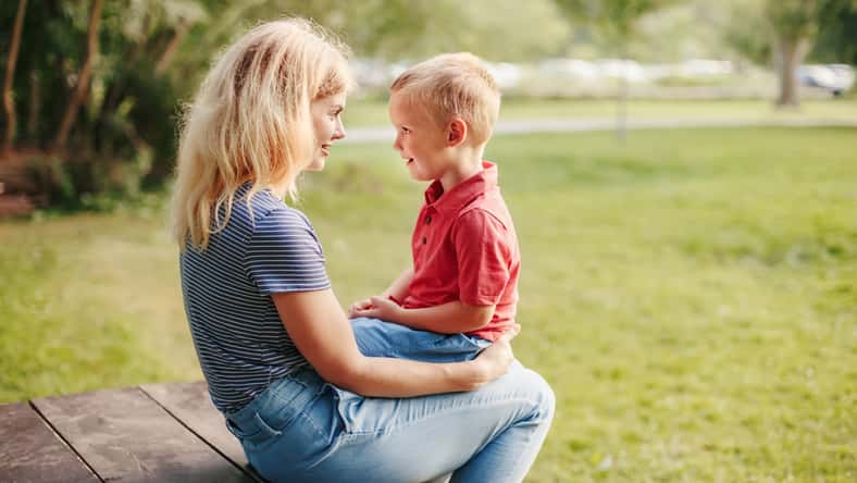 Young Caucasian mother and boy toddler son sitting together face to face. Family mom and child talking communicating outdoor on summer day. Happy authentic family childhood lifestyle.