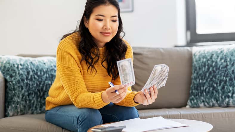 accounting, taxes and finances concept - young asian woman with papers and calculator counting money at home