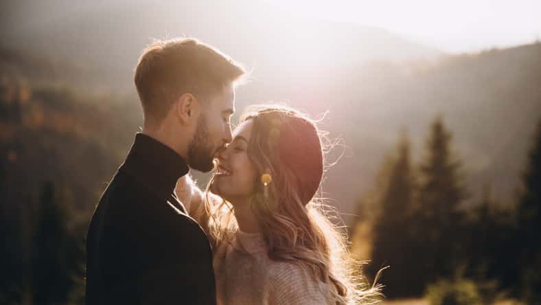 Stylish model couple in the autumn mountains. A young guy and a girl hug and kiss on a background of a forest and mountain peaks at sunset.