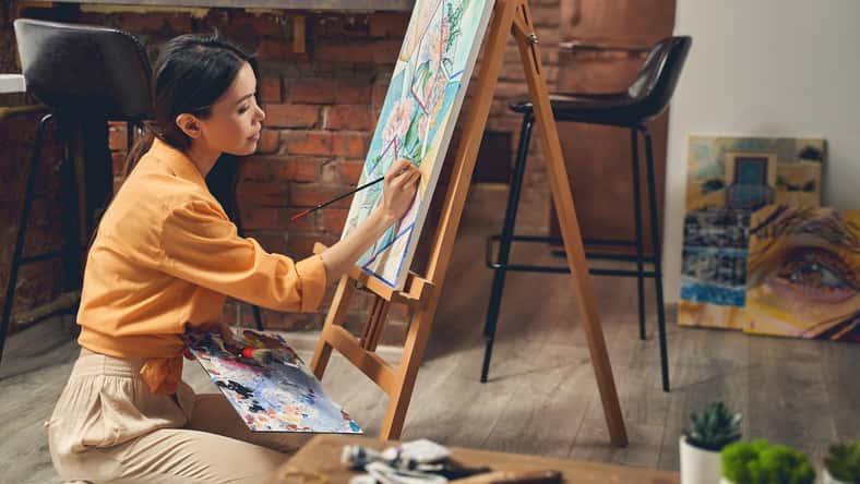 Talented female artist sitting on the floor and drawing flowers with paints on easel in home art studio