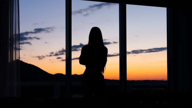 Silhouette of a young woman standing in front of a window with sunset in the background