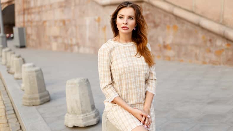 Young elegant woman stays alone in the city, dressed in a classic dress with straight and red long hair.