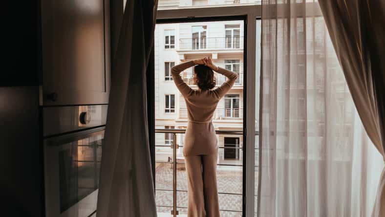 Photo from back of woman in brown pants. Indoor shot of caucasian female model looking at city through the window.