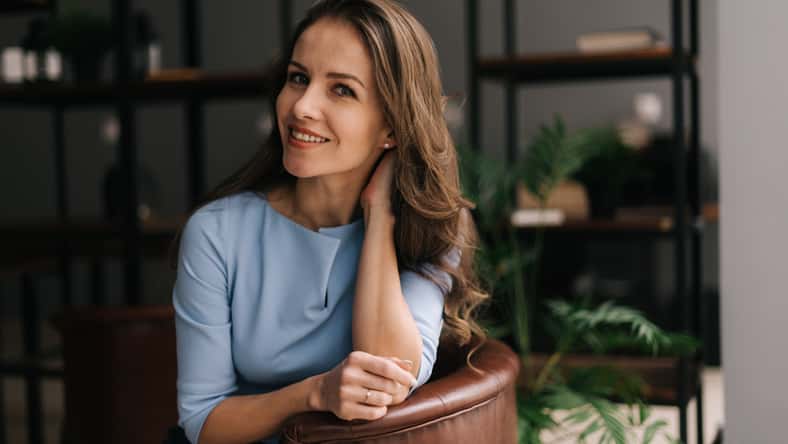 Attractive young businesswoman wearing stylish light blue dress sitting at the chair in office with modern interior, looking at the camera. Portrait of charming business lady.