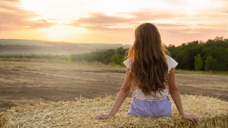 Adorable little girl child sitting on a hay rolls in a wheat field at sunset back view