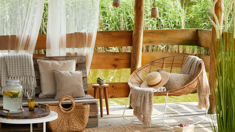 Interior design of summer gazebo by the lake with stylish rattan armchair, coffee table, sofa, pillows, plaid and elegant accessories in modern decor. Summer vibes.