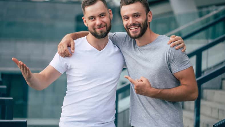 Two happy excited handsome bearded best friends in casual clothes are handshaking, hugging while standing outdoor.