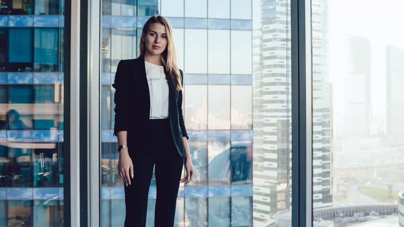 Half length portrait of pretty blonde business woman in trendy elegant formal wear standing near window in office interior, confident beautiful 30s female executive manager in suit looking at camera