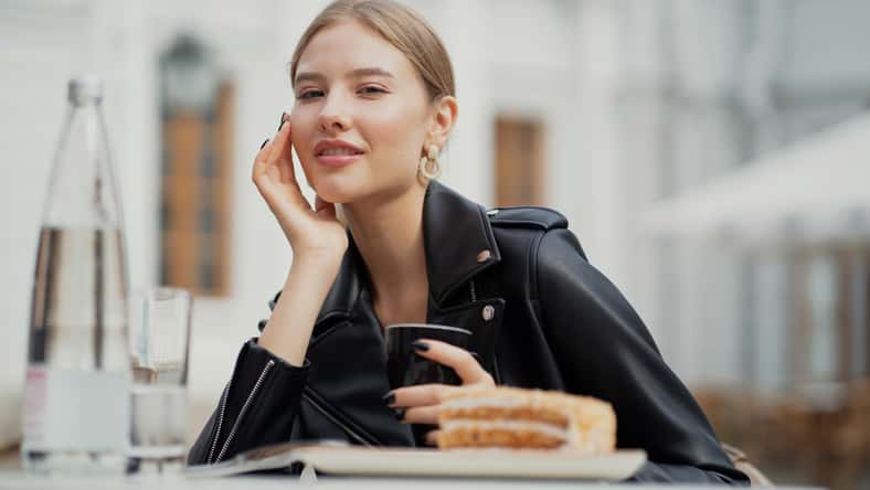 enjoying a delicious coffee, a young woman of European appearance ordered Breakfast, a Cup of Americano and delicious sugar desserts cake. black leather jacket, straight blonde hair.