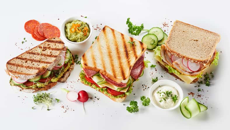 Trio of gourmet sandwiches on assorted bread with meat, cheese, guacamole and pesto fillings surrounded by ingredients and seasoning on white in a panorama banner