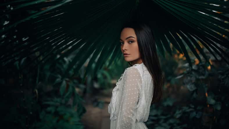 Portrait of young woman under big leaf of palm tree inside orangery.