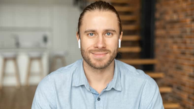 Portrait of an attractive young bearded hipster man smiling at the camera, wearing headphones, standing in the modern house, wearing a casual shirt. The view while the video-calling concept