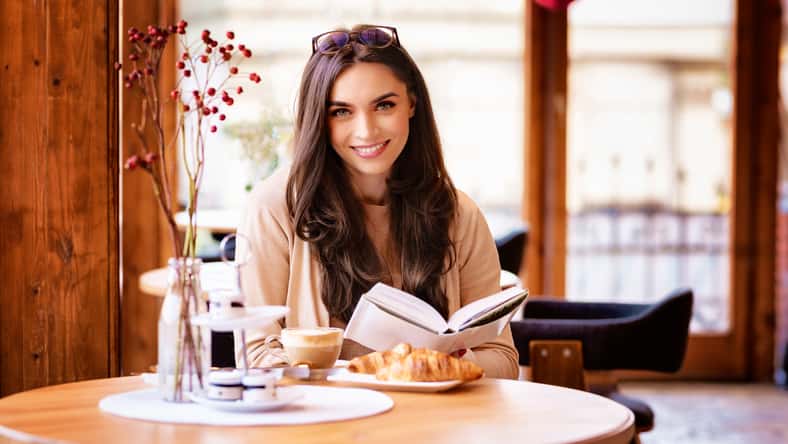 Portrait of beautiful young woman having breakfast and reading book while sitting at cafe desk.