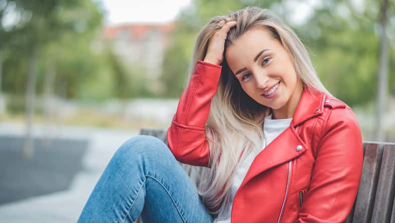 Smile young beautiful woman sitting on bench. Outdoors daylight. Smiling pretty girl wearing casual clothing. Outside